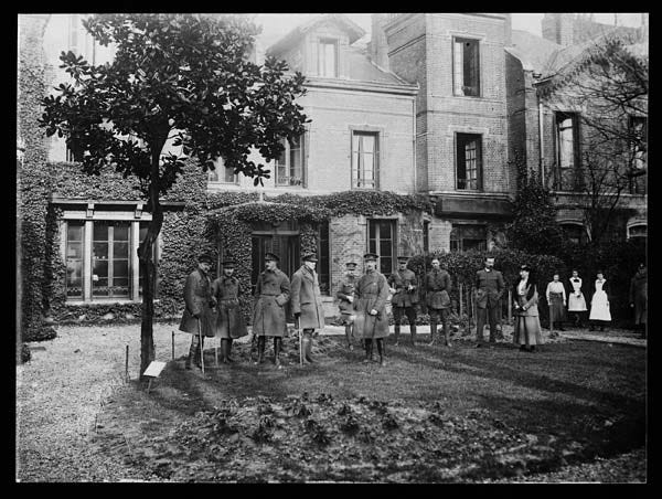 A group of officers stand outside a rather nice, ivy-covered, building. They are looking at the camera and are, quite frankly, looking a bit awkward. In the background some men and women without uniforms stand. They are the staff of the club. The officer sixth from the left looks a lot like Charlie Chaplin.