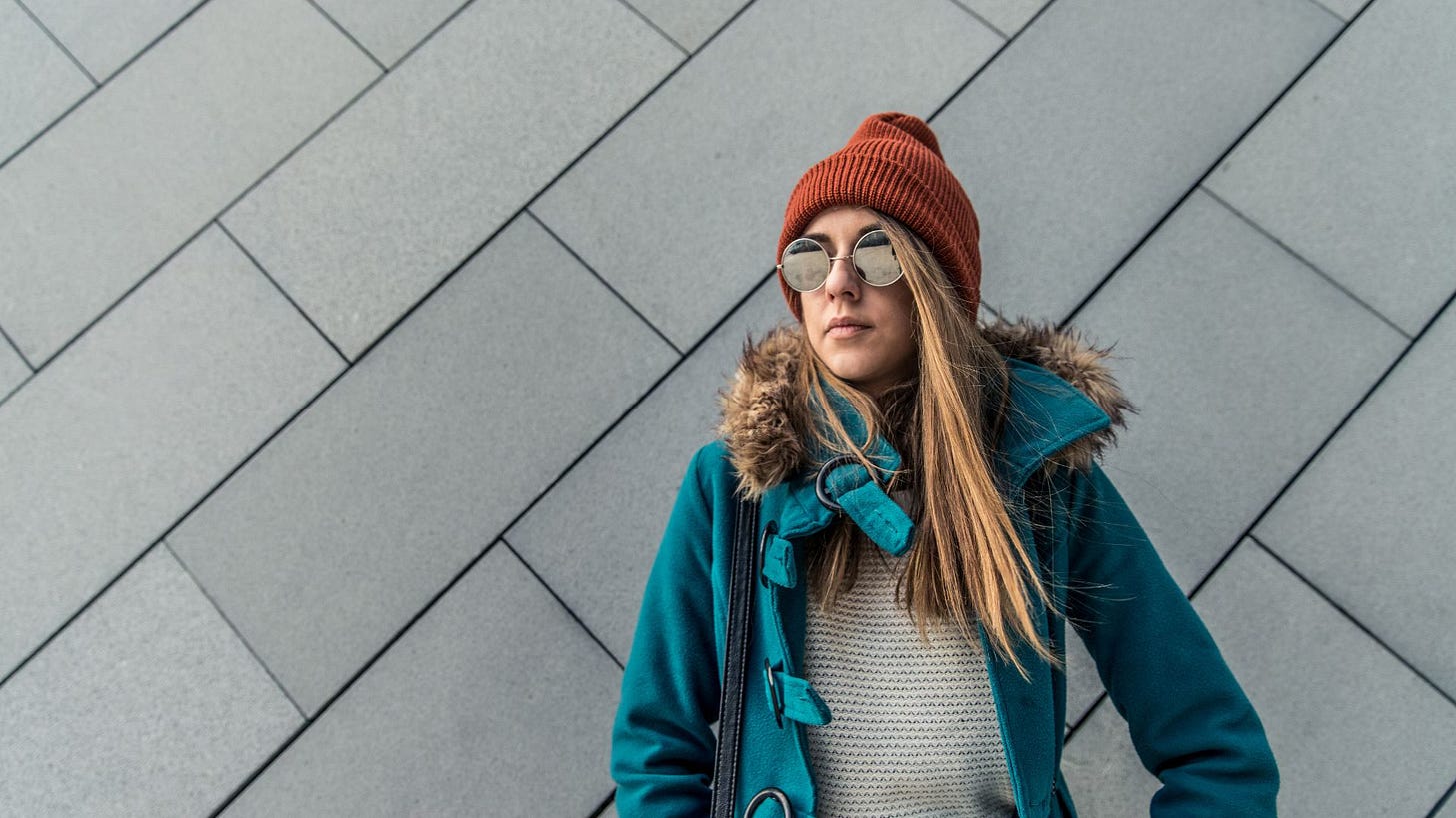 White woman with long hair wears a teal coat and orange beanie and sunglasses. Photo credit: Canva.