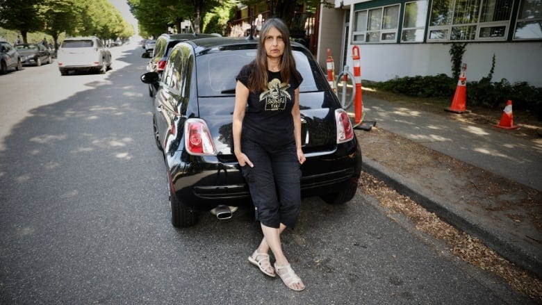 Teresa Chhina sits on the rear bumper of a black Fiat car on a street in Vancouver.