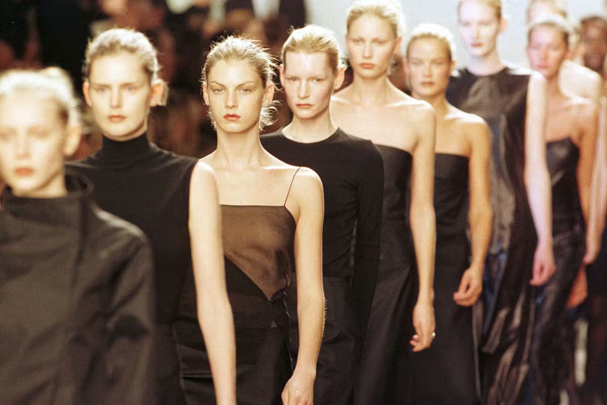 A row of models seen from a three-quarter view wear a selection of darkly colored clothes as they walk the runway in single file. 