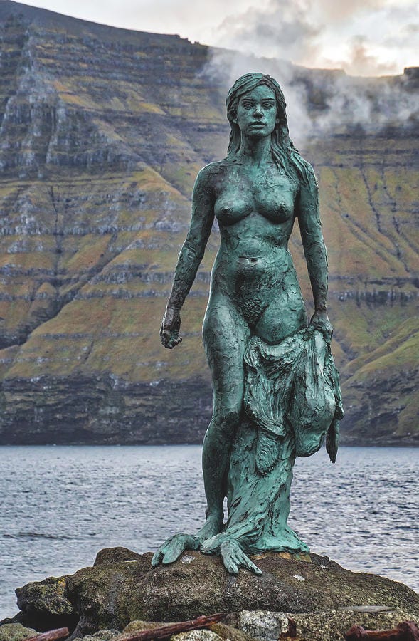 Statue in verdigris of a naked seal women walking out of the sea carrying her seal skin with a background of a cliff on the Faro Islands