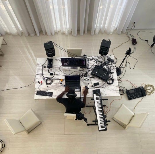 A black person at a white table in a white room surrounded by electronics.