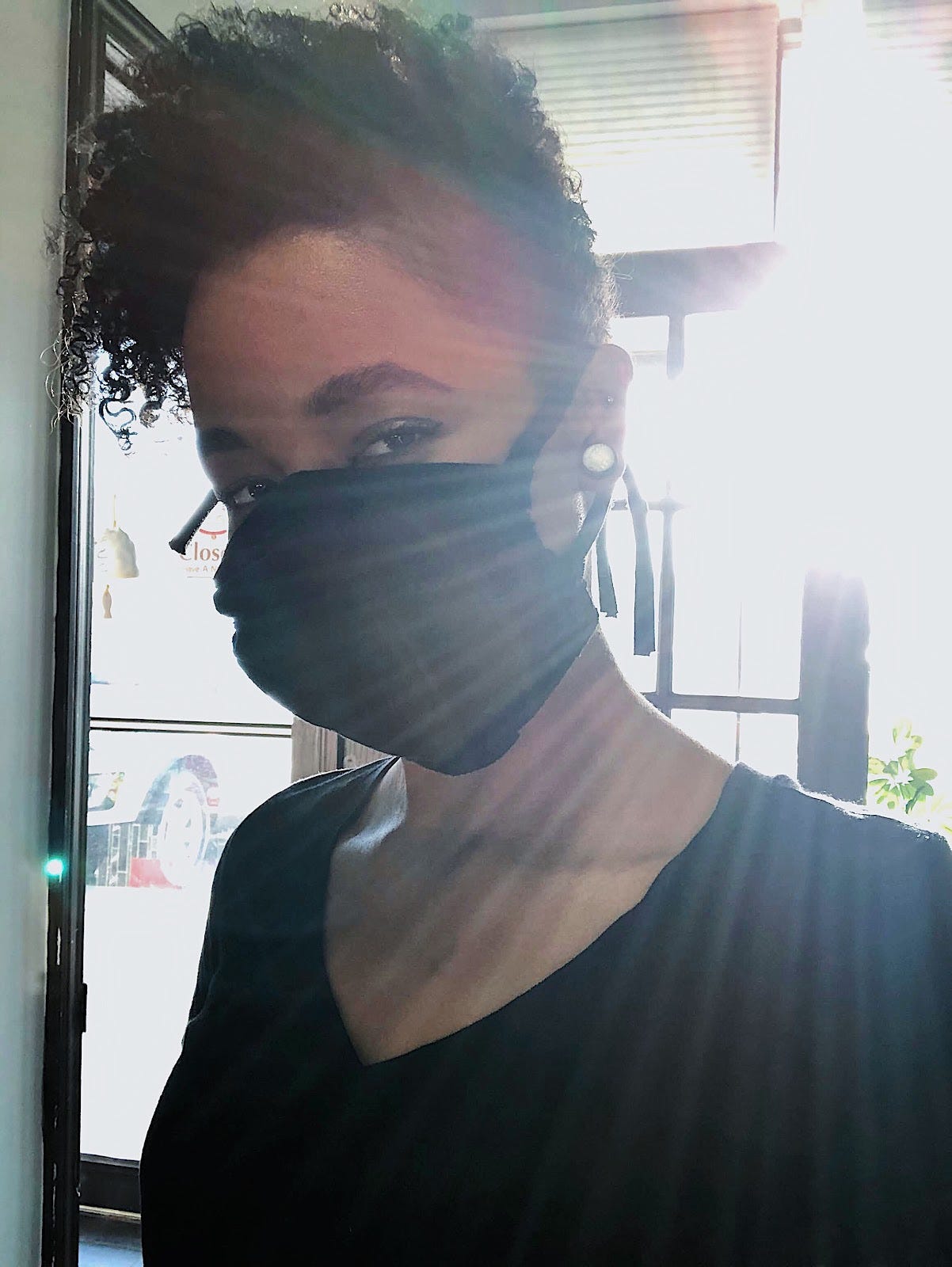 A black girl with a black mask on smiling with her eyes at the camera while waiting by the door of a restaurant, showing off her cut, curly hair