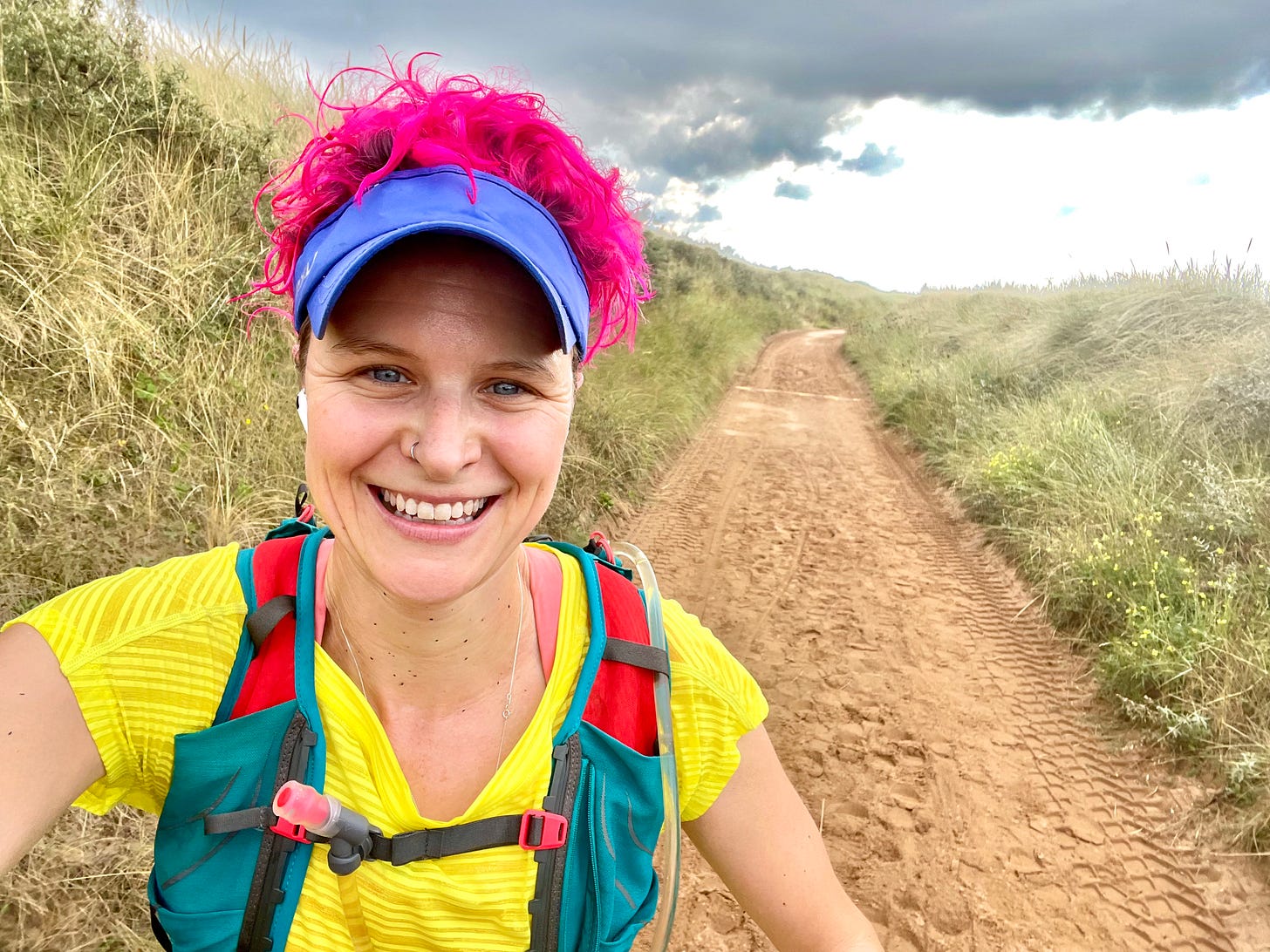 Anna McNuff, mid-run on a sandy trail, grinning into the camera. She wears a bright yellow top, purple visor, and turquoise hydration vest, with vivid pink curls peeking out. The path stretches behind her through tall grass under a moody sky.