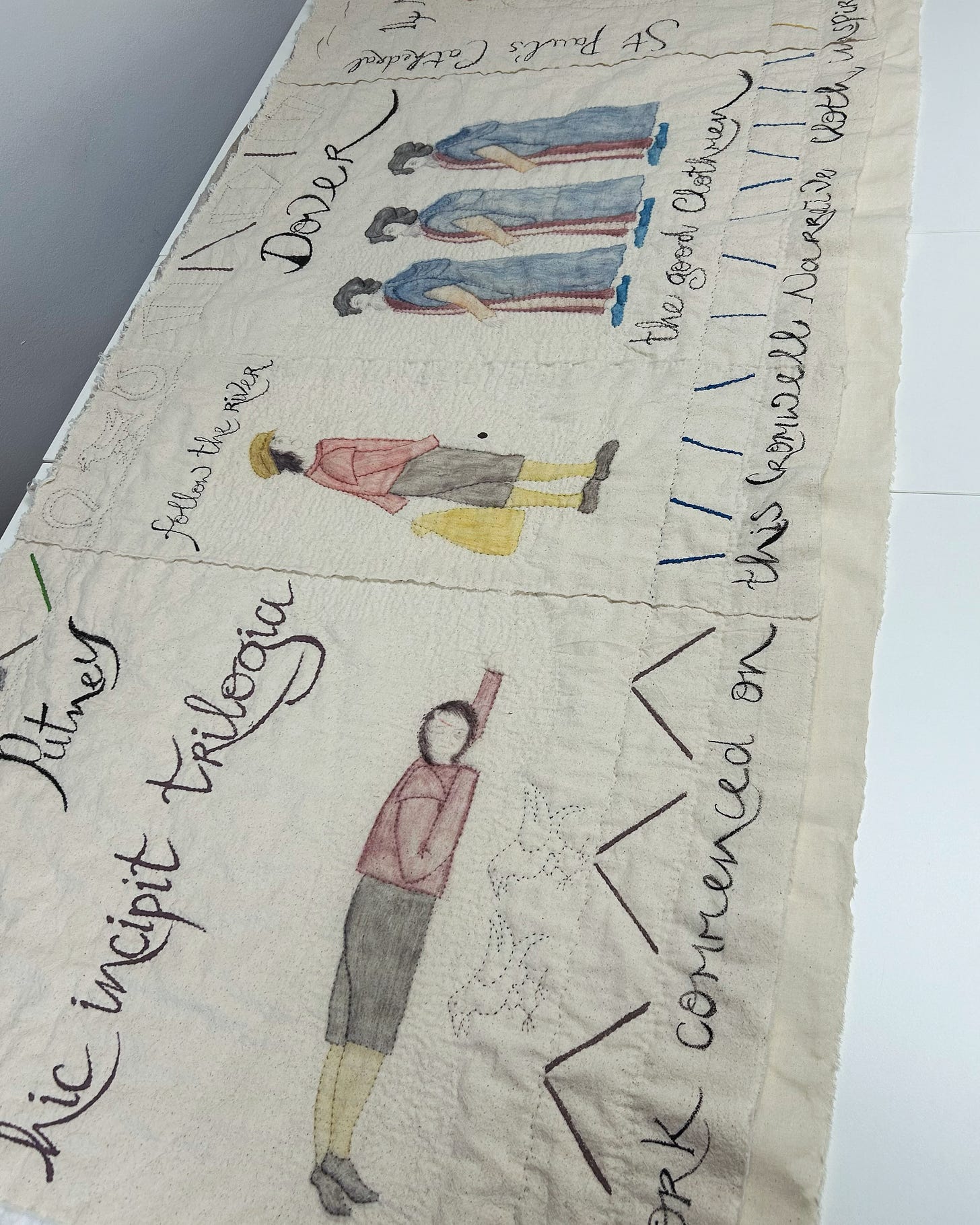 A long piece of cream fabric with figures painted and quilted on it, along with dates and places. The piece shows a boy lying on the ground; a boy walking; three older men with beards. 