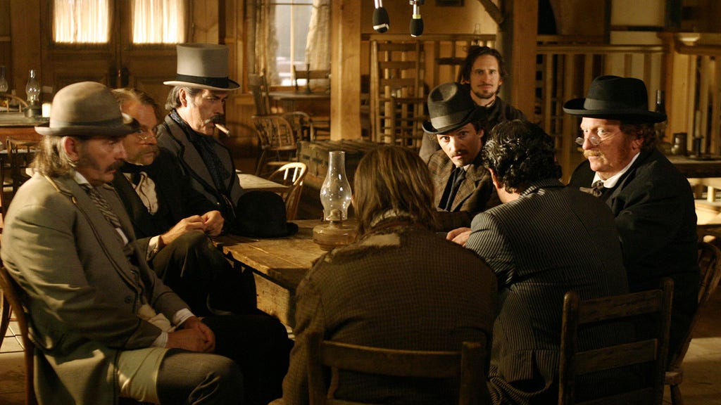 This image shows the town council assembled by Al Swearengen in "Plague," Deadwood's sixth episode. From left to right: Tom Nuttall (played by Leon Rippy), Doc Amos Cochran (played by Brad Dourif), Cy Tolliver (played by Powers Boothe), Reverend H.W. Smith (played by Ray McKinnon), hardware merchant Sol Star (played by John Hawkes), publisher A.W. Merrick (played by Jeffrey Jones), Swearengen (played by Ian McShane, with his back to camera), and hotelier E.B. Farnum (played by William Sanderson, also with his back to camera).