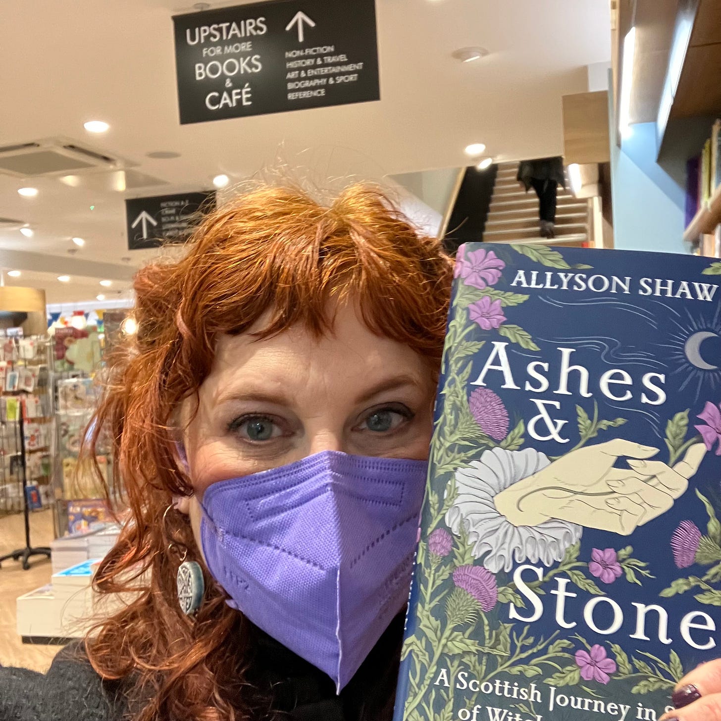 A redheaded woman in a mask--that's me--holding a copy of her book Ashes and Stones in a big chain bookstore