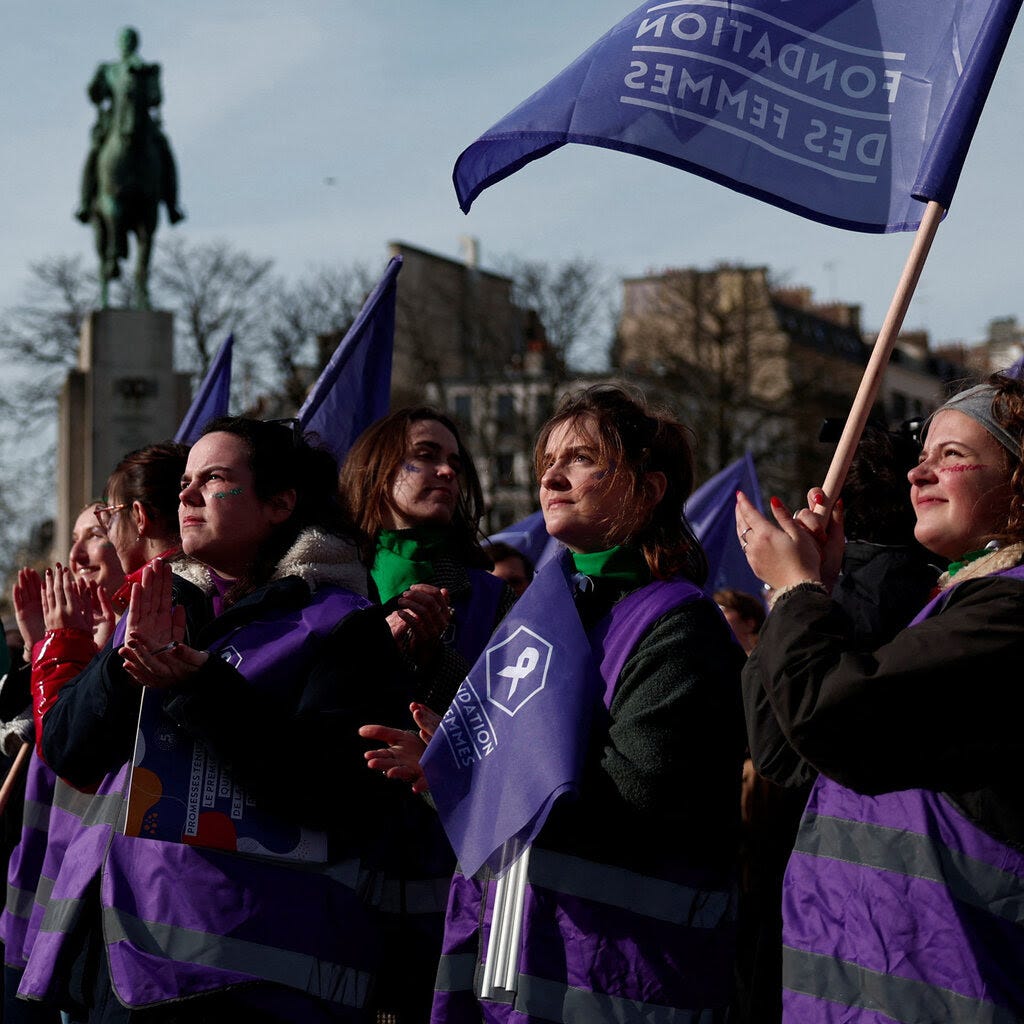 A group of people in purple vests and green scarves stand together waving purple flags.