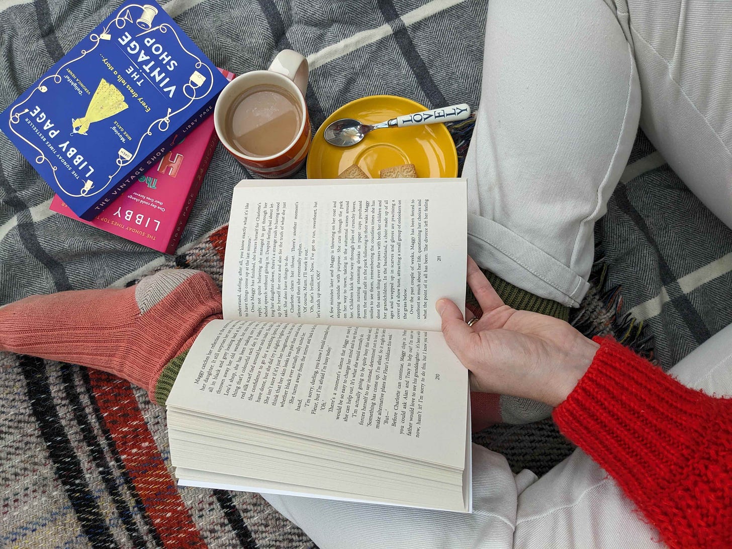 A crop of someone sat on blankets reading a book with a mug of tea and biscuits next to them.