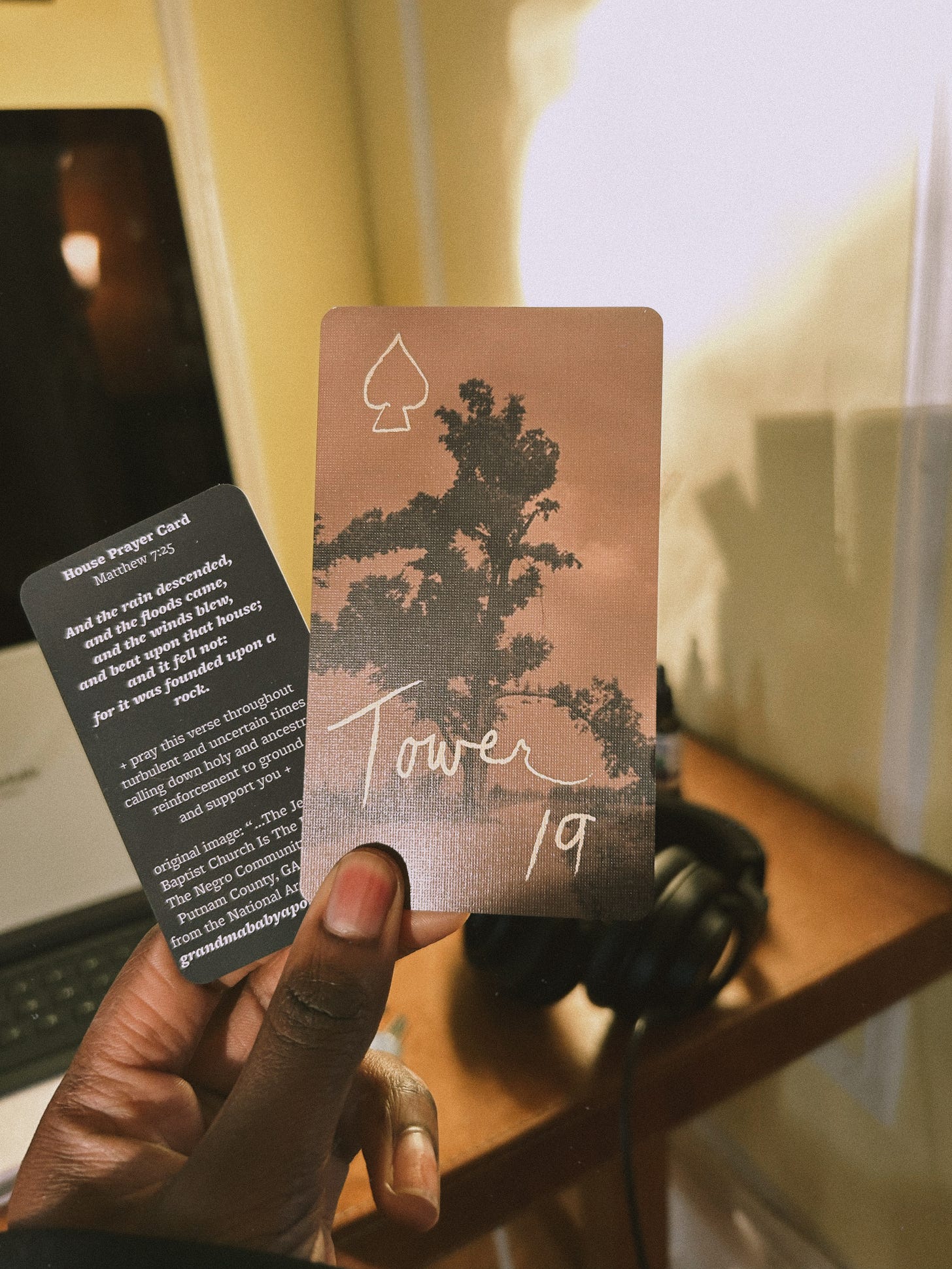 Ismatu holds the card they drew for this essay in the new morning light. There are two cards in their hand: one that is smaller, black, and quotes Matthew 7:25 and another larger card with a tree in sepia tones, a drawn spade, and a handwritten note: Tower