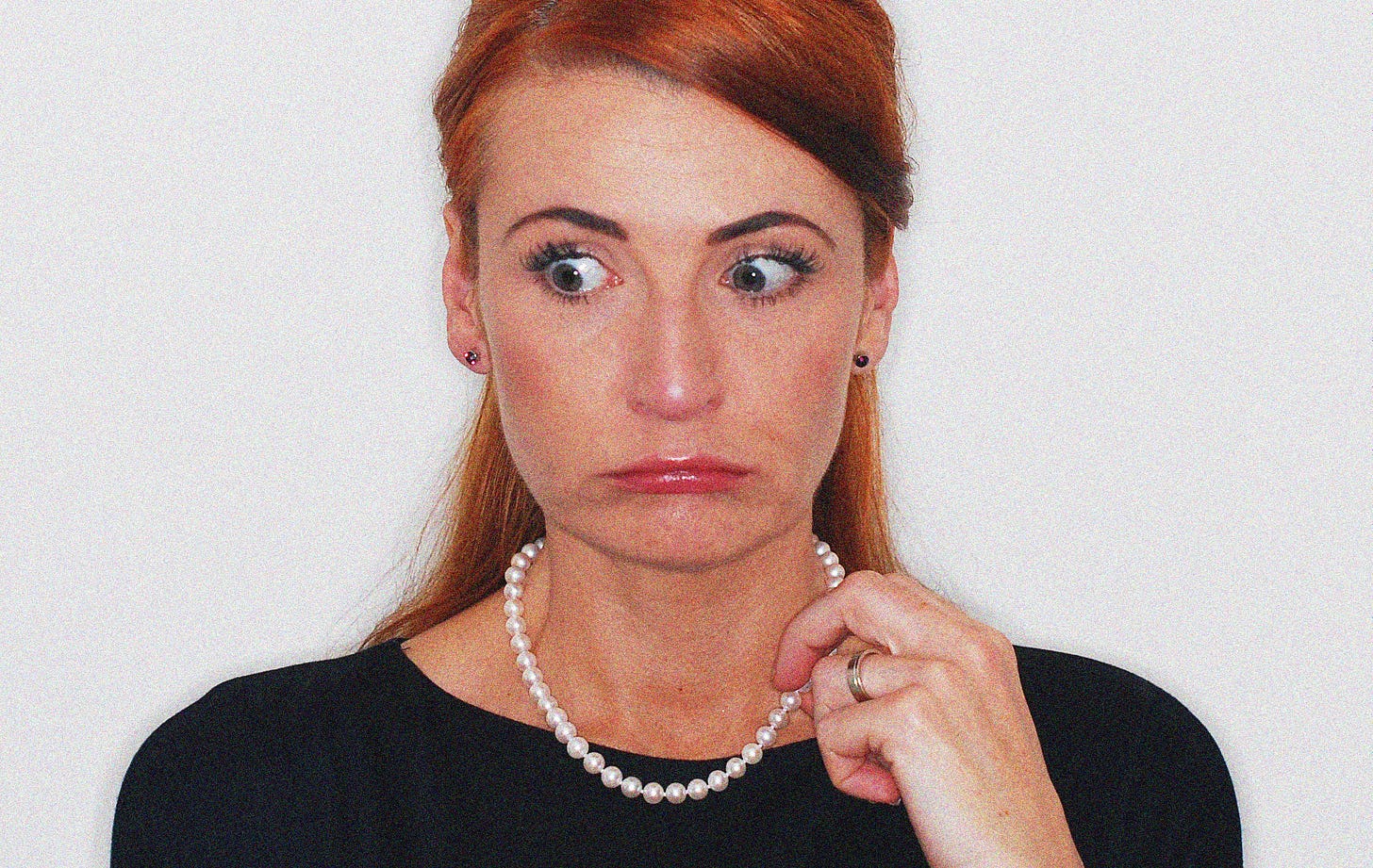 A head and shoulders shot of Catherine, a (then) 42 year old white woman with long red hair, her eyes wide and shocked and mouth closed with lips and cheeks puffed out. She is clutching a string of pearls with one hand and wearing a plain black top