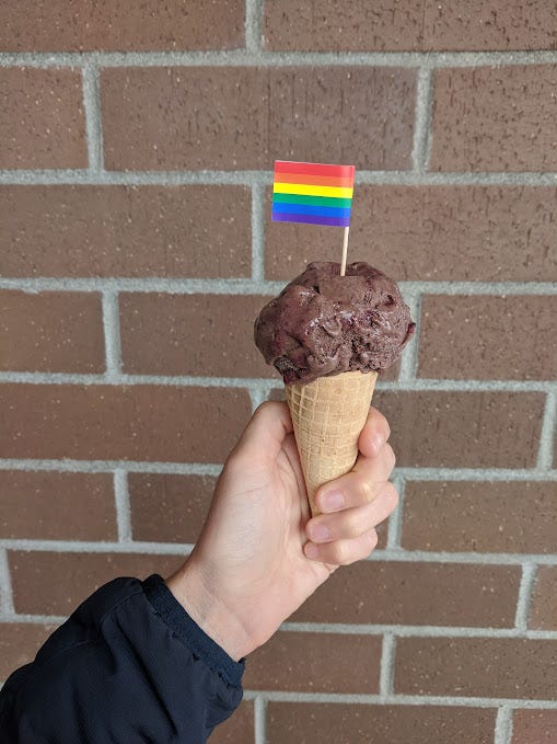 a pale arm holds up a cone of chocolate ice cream with a small rainbow flag on top