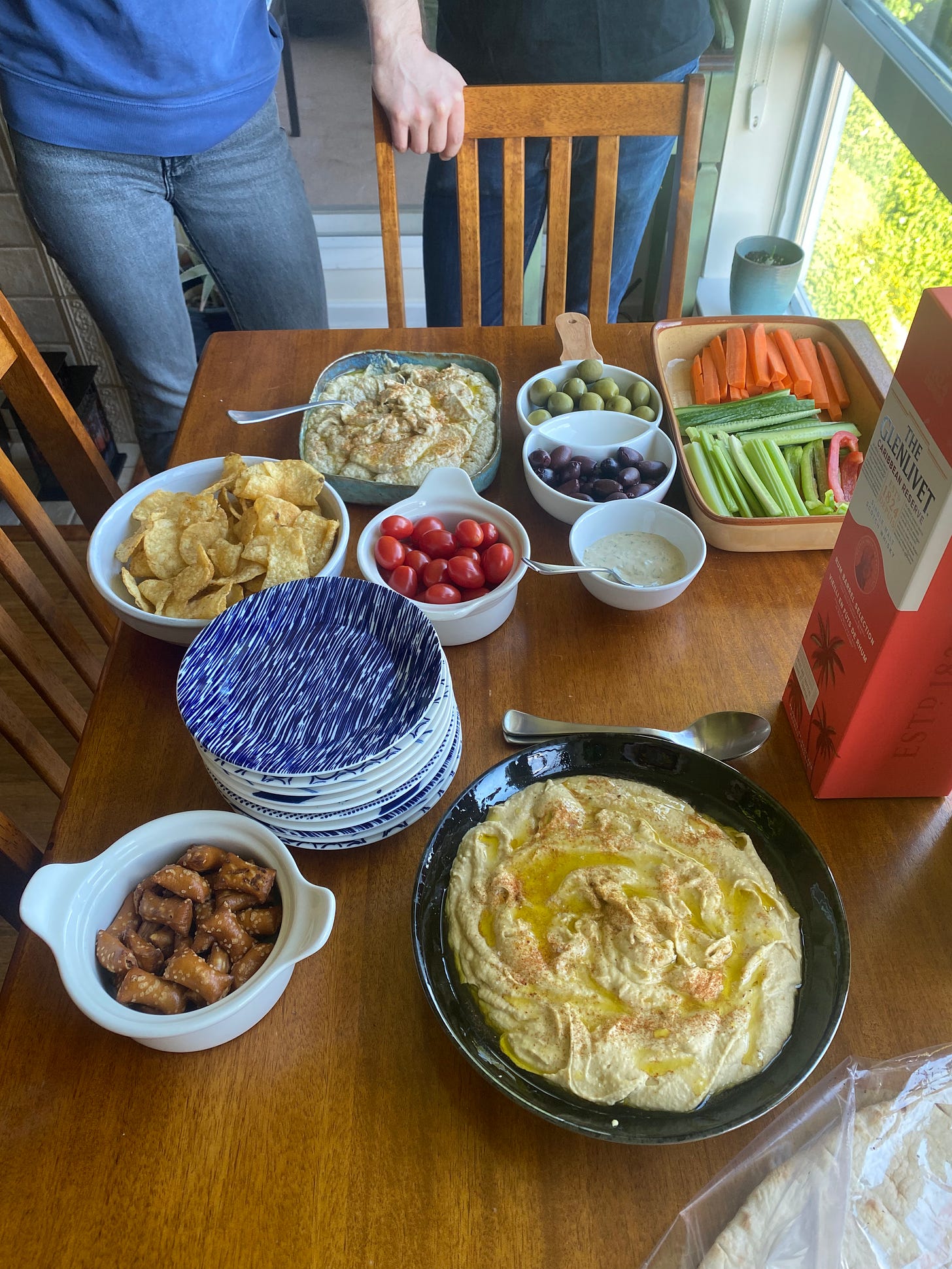 A spread of hummus, baba ganoush, vegetables, olives, chips, and pretzels, with a stack of small plates. Just visible in the corner is a bag of flatbread, and a box containing a bottle of Glenlivet scotch.