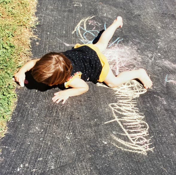 A three-year-old child wearing a polka-dot top and yellow shorts lies on their stomach on a black piece of pavement. On the pavement is an exuberant scribble done in light-colored chalk. She holds a piece of chalk in her hand right hand, which she is using to make more marks.