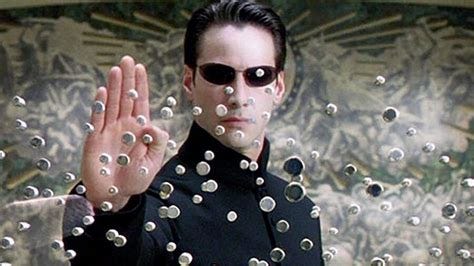 Keanu Reeves as Neo in the Matrix, black suit and sunglasses holding up his hand to stop lots of bullets