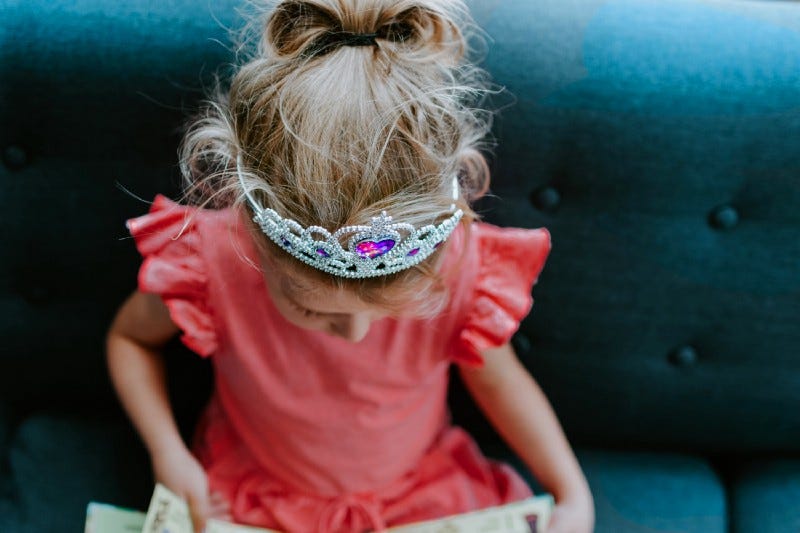 Little girl on a sofa wearing plastic crown while reading.