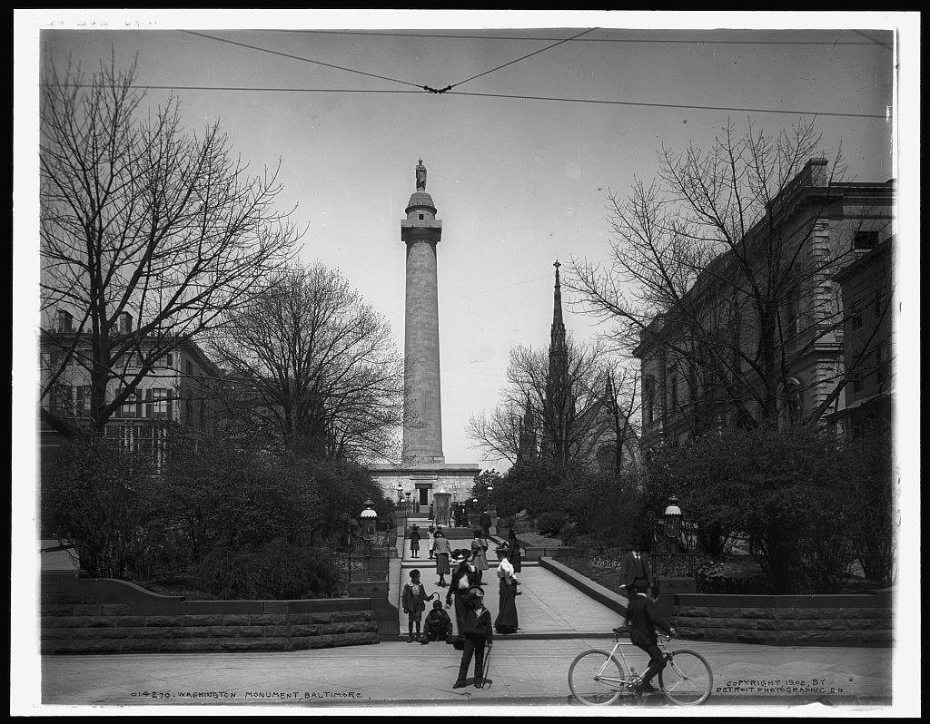 Black-and-white photograph depicting Baltimore’s Washington Monument from the street during the winter. Pedestrians and a cyclist are passing by and some are admiring the monument.