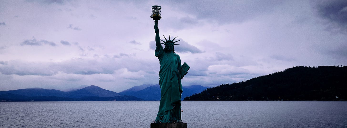 A dark photograph of a replica of the statue of liberty with a large lake and mountains in the background.