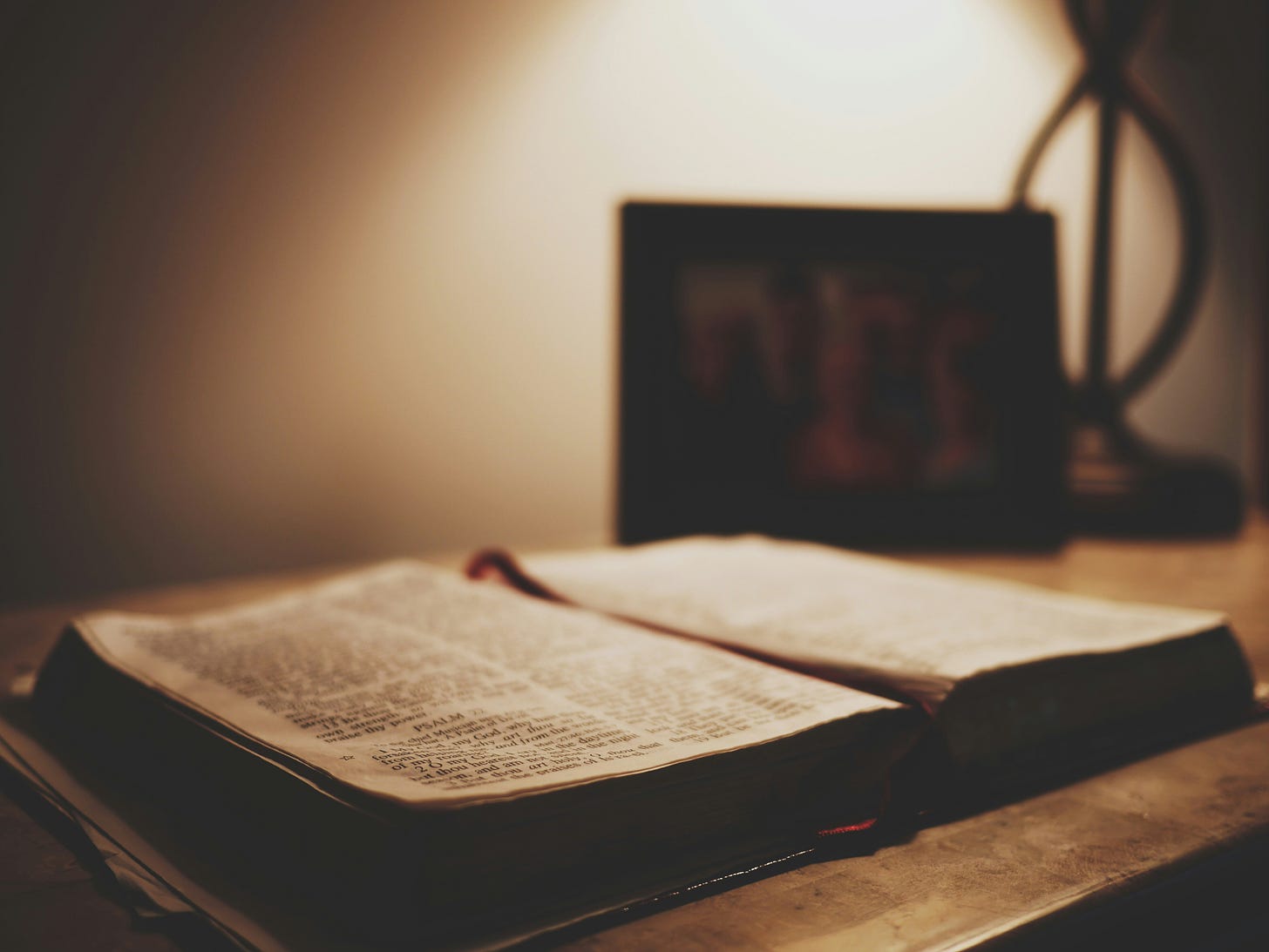 Image of an open Bible on a desk with a framed picture under a lamp.