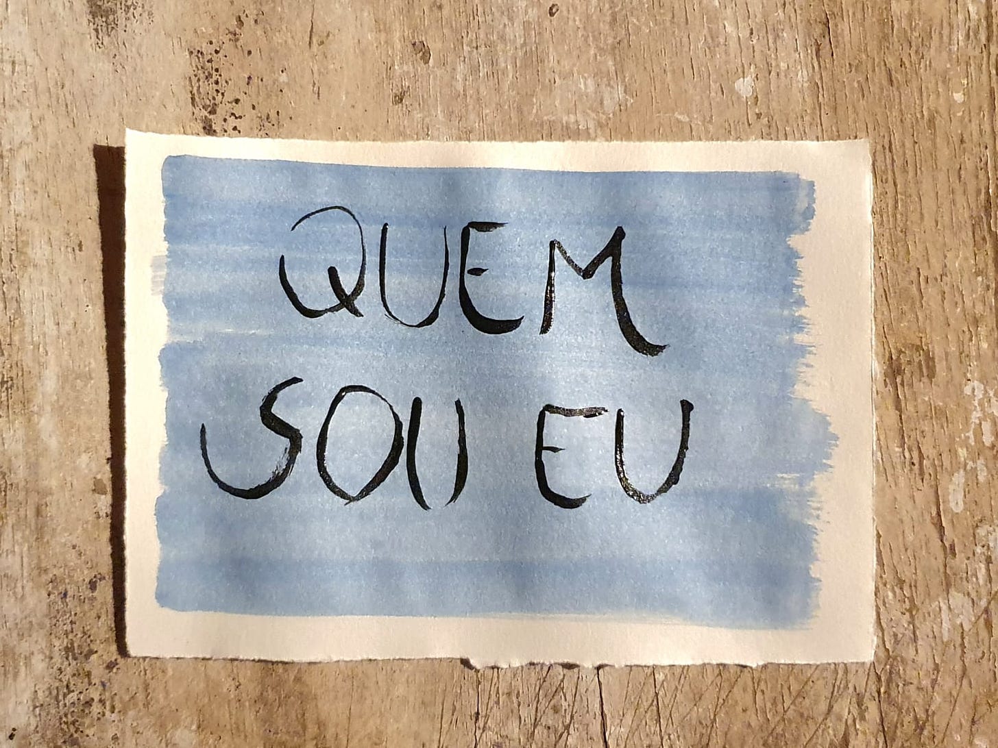 Fotografia de um papel pintado de azul sobre um fundo de madera com o escrito "Quem Sou Eu"