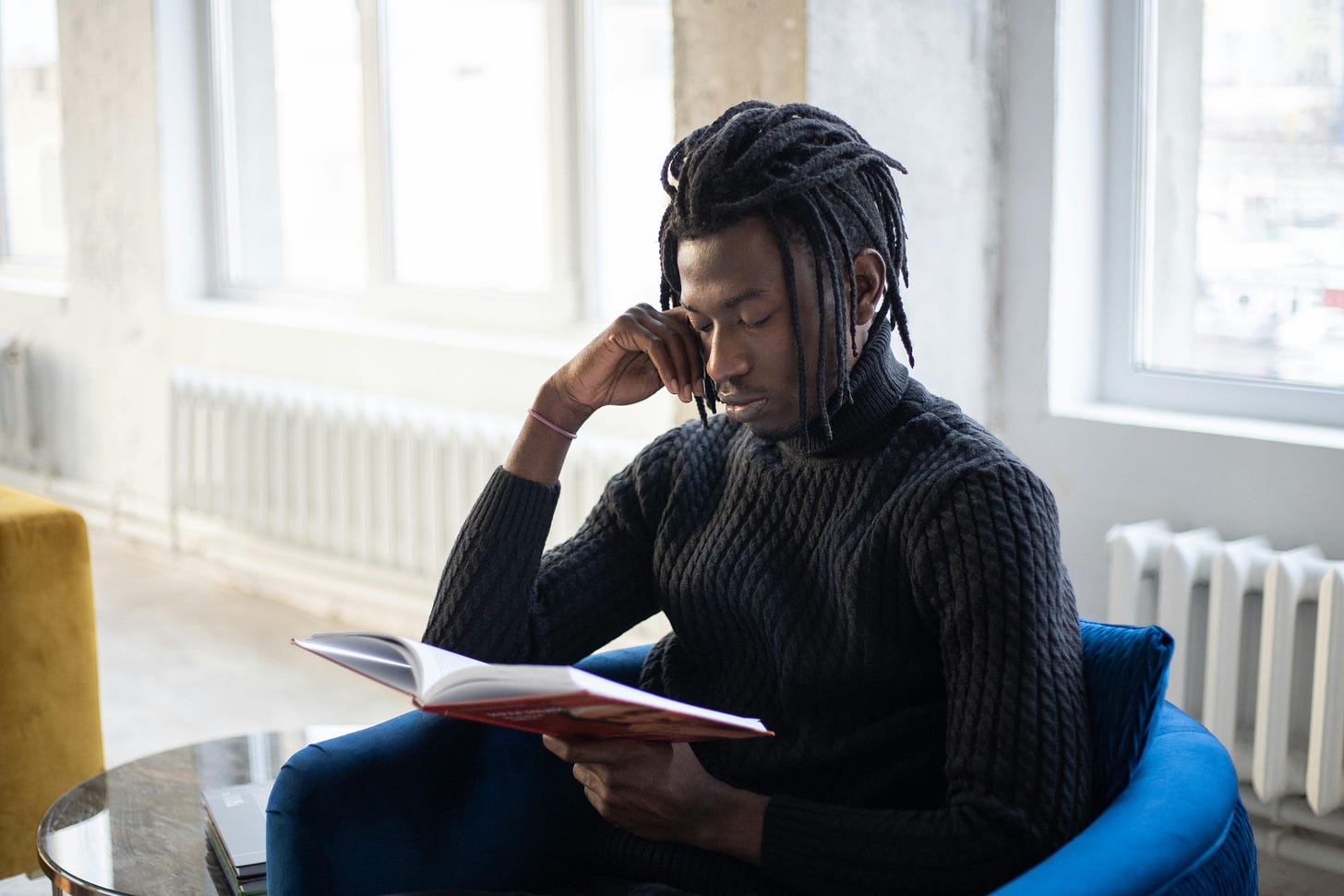 A young Black man with 'locs sitting in a chair and reading a book.