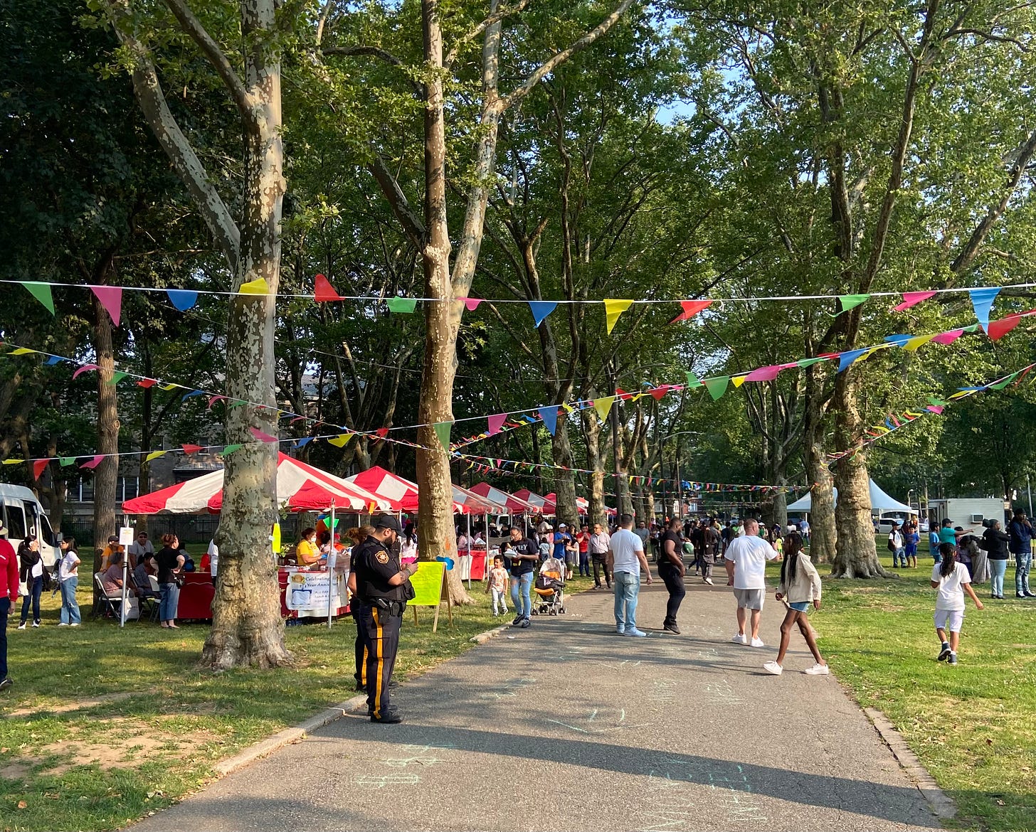 Pic of Lincoln Park with the carnival in it -- people milling about, trees, grass, a stream of those brightly colored triangle flags hanging over the pathway; a row of tables with vendors with red and white umbrellas.