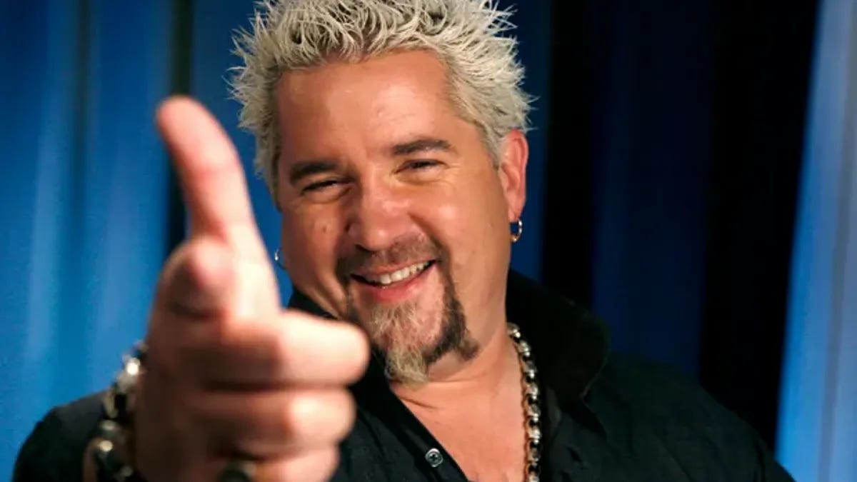 Guy Fieri in front of a blue curtain, smiling, pointing directly at the camera with a finger gun