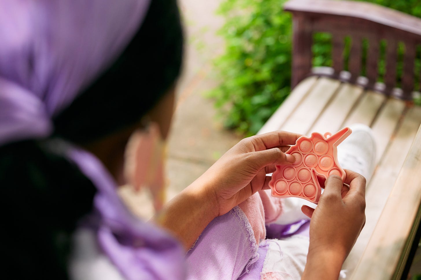 Una donna nera autistica viene mostrata di spalle mentre usa il suo stim toy popsocket rosa. La donna ha lunghi capelli ondulati legati all'indietro con un foulard color lavanda e siede su una panchina al parco.