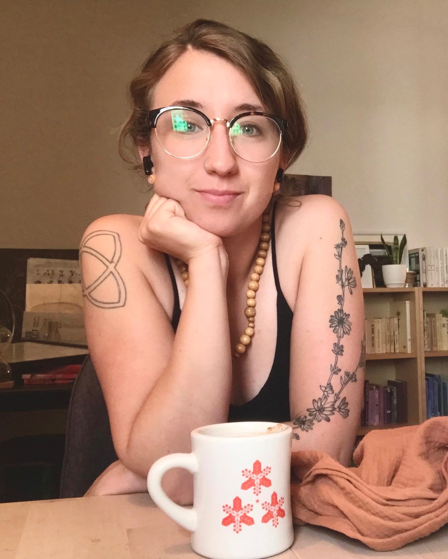 a white woman with brown hair and glasses is resting her chin on her hand. she is slightly smiling at the camera. she has several tattoos on her arms. 