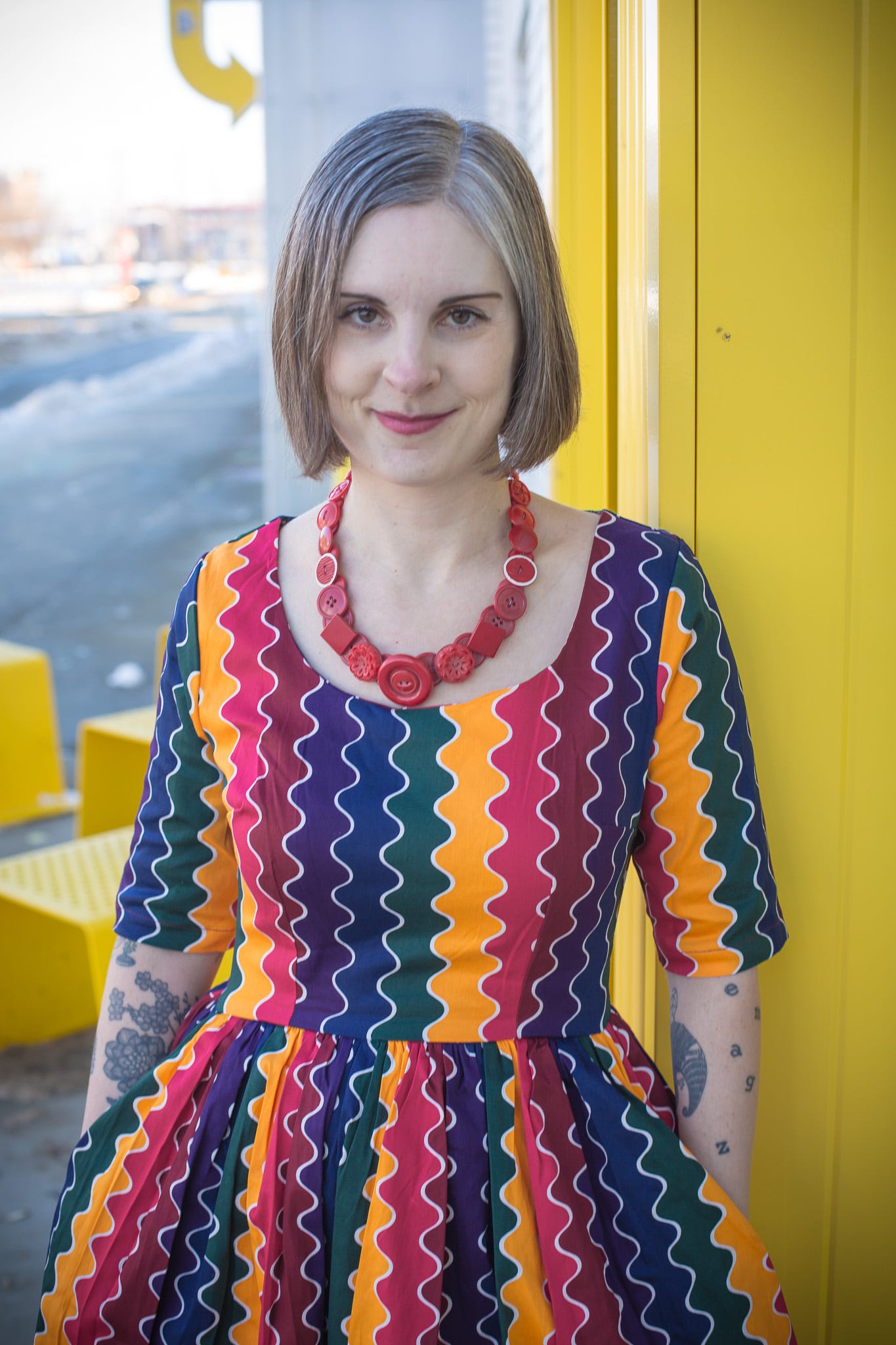 Photo of Jen outside of co-lab in downtown Edmonton, December 2022. She's wearing a squiggly rainbow dress and red button necklace. 