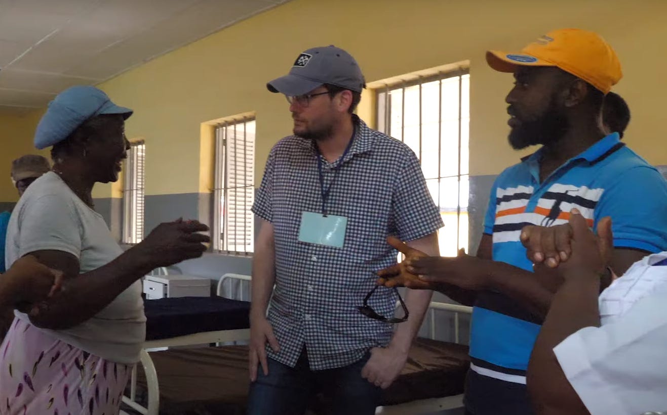Three people stand and talk inside a room with hospital beds and barred windows, engaged in conversation.