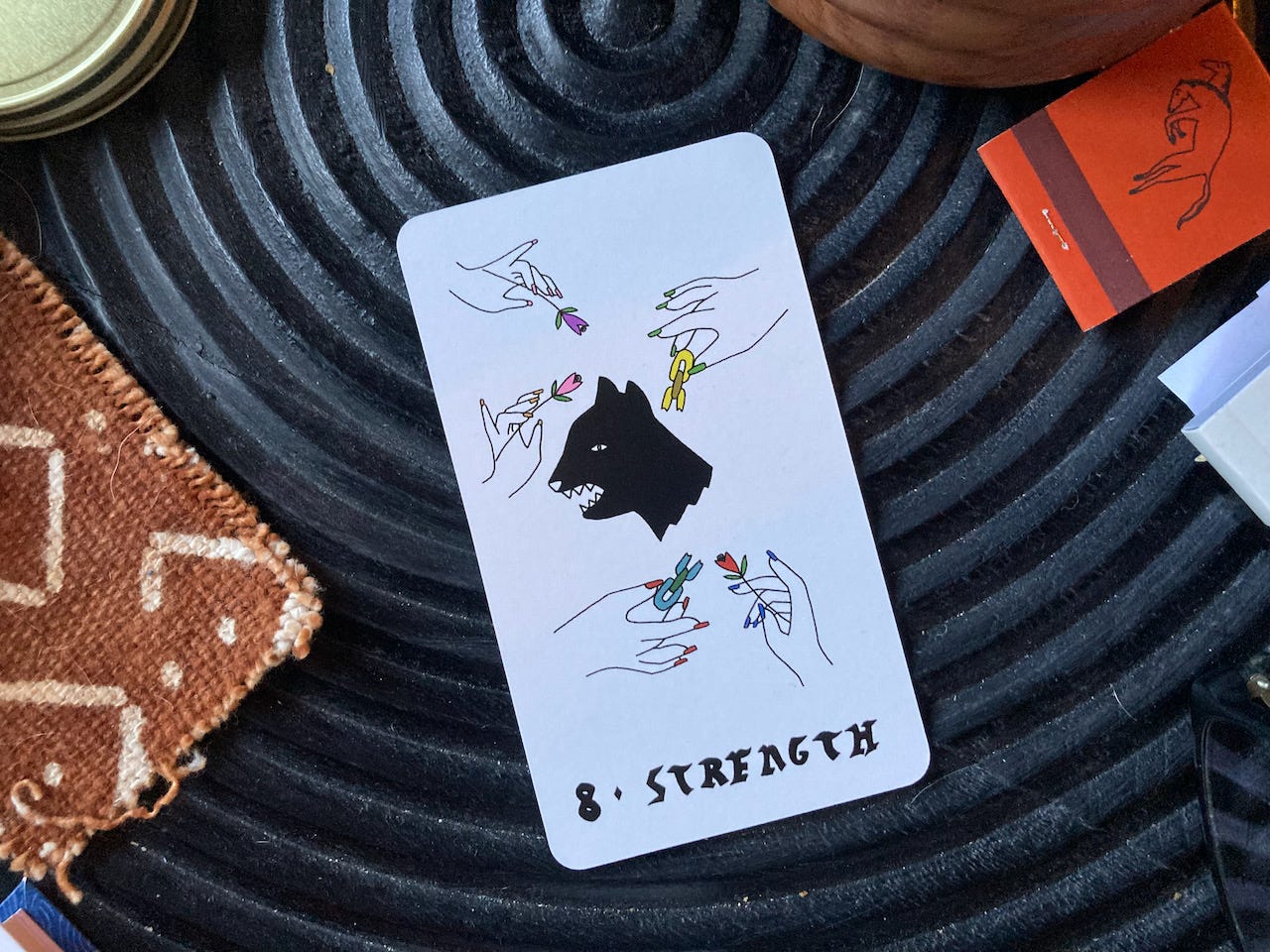 A Strength tarot card rests on a black table. The card shows a folksy drawing of a black cat or panther snarling while hands surround it holding flowers and broken chain links.