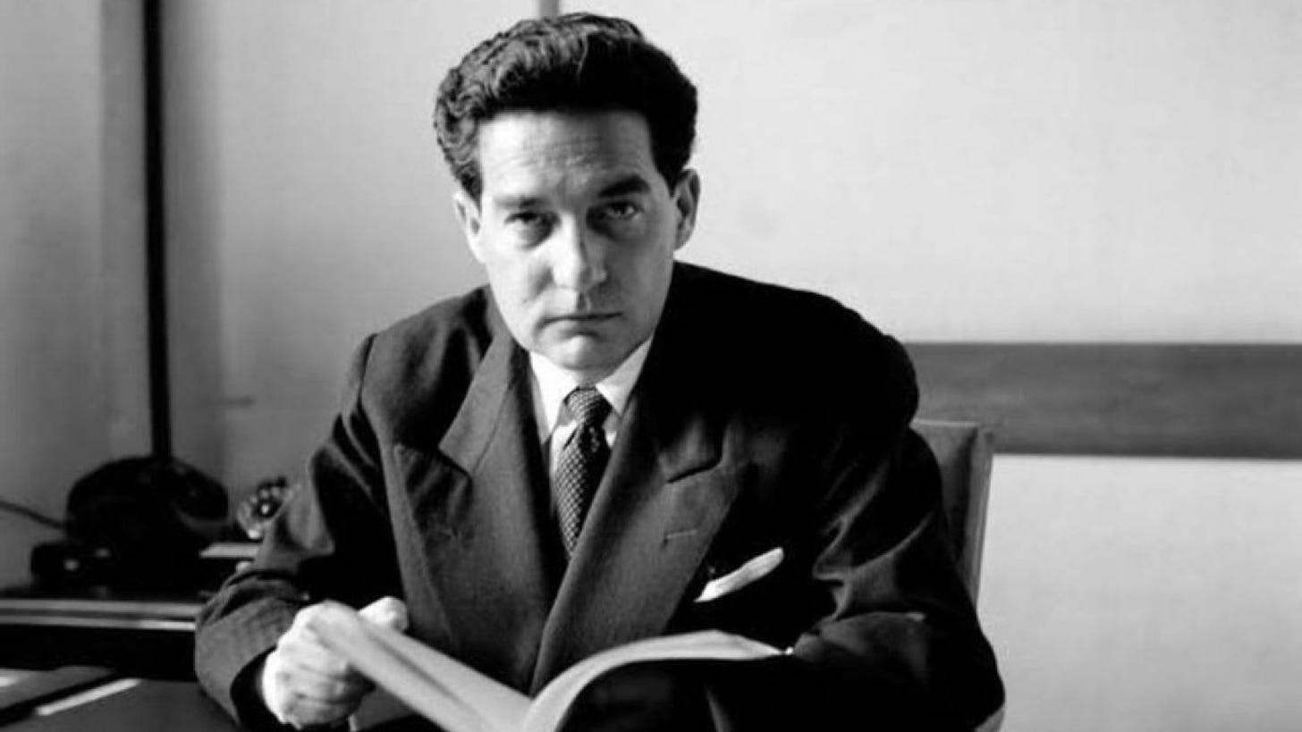 Black and white photo of a middle-aged man with dark hair. He is holding open a book and looking directly at the camera. He wears a suit.