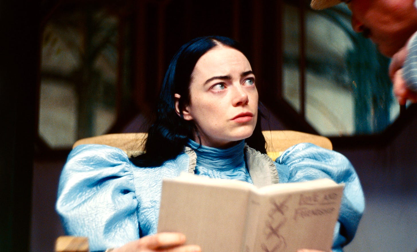 A woman in a blue dress with puffy sleeves looks up from the book she is reading