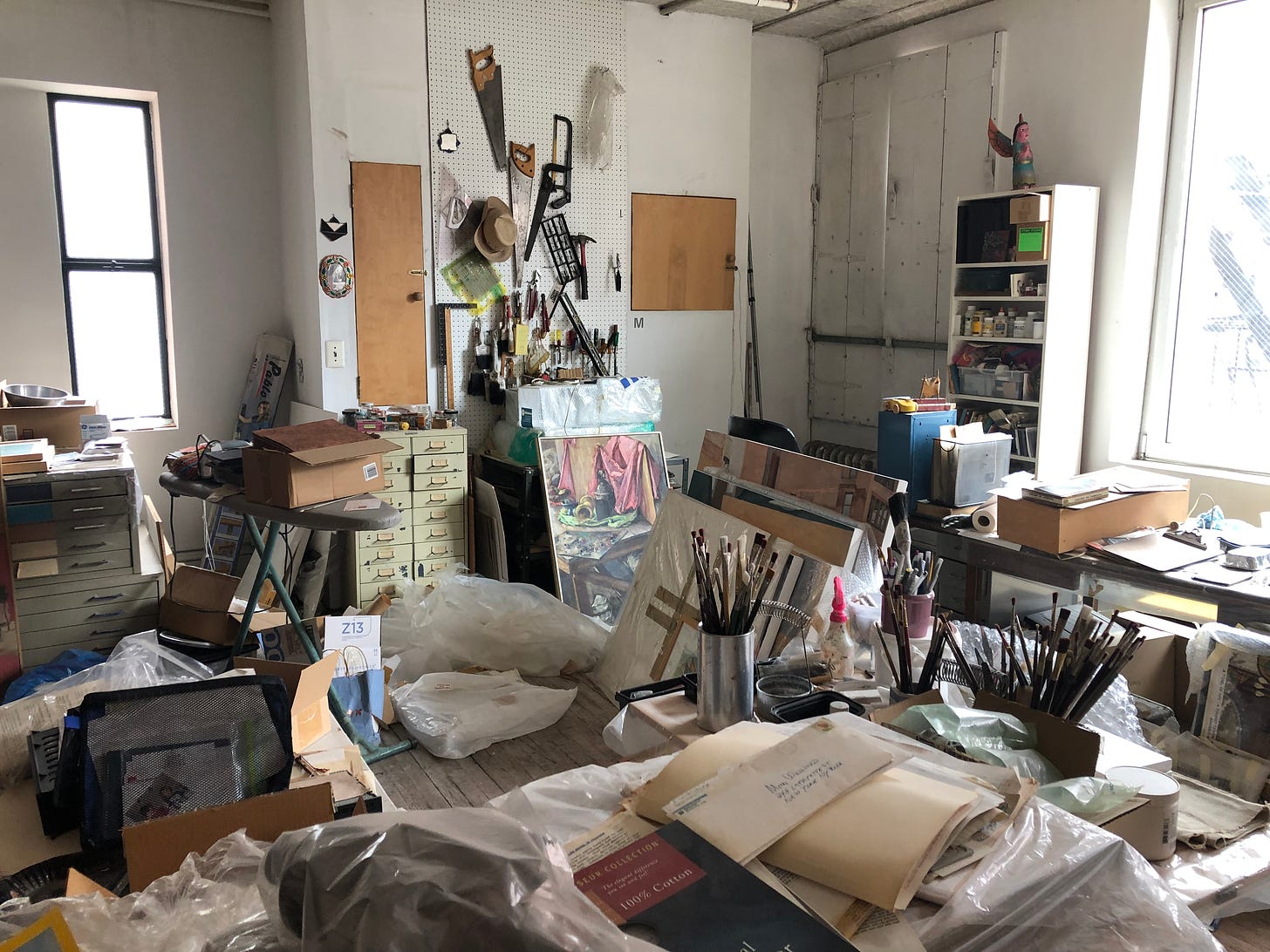 A corner of an art studio full of art,  tools, supplies, paintings, in a jumbled mess.
