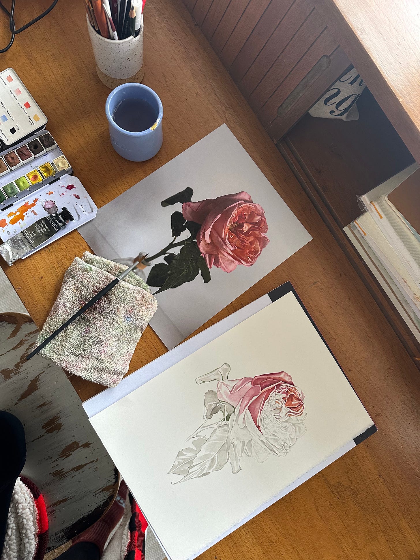 A birds eye view looking down at a wooden desk space with a drawing half painted of a single pink rose, with a reference photo laying next to it, and watercolors and paintbrushes laying to the left of it