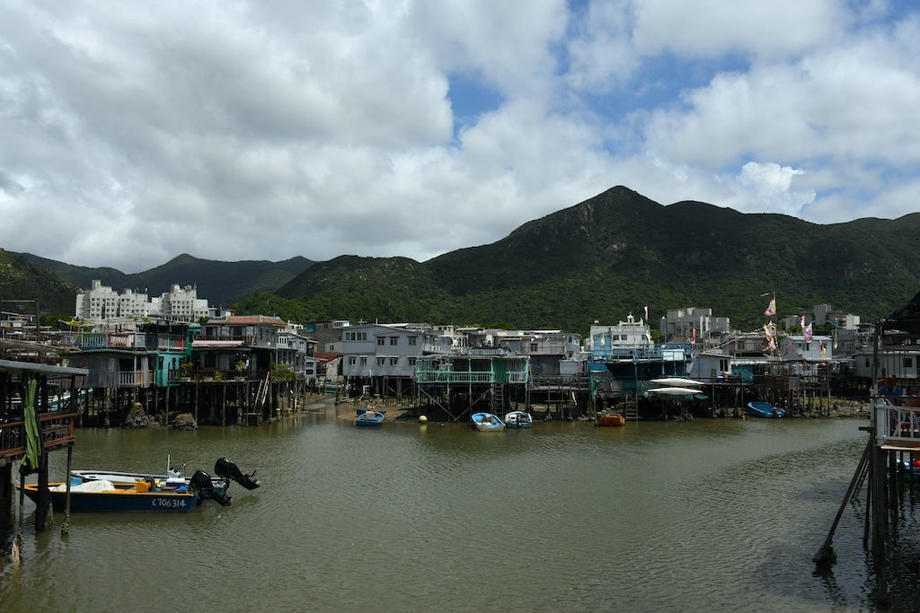 Foto a colori di uno scorcio di Tai O. Si vede il fiume in primo piano, con le palafitte tutte diverse tra loro. Sullo sfondo, le montagne e il cielo nuvoloso.