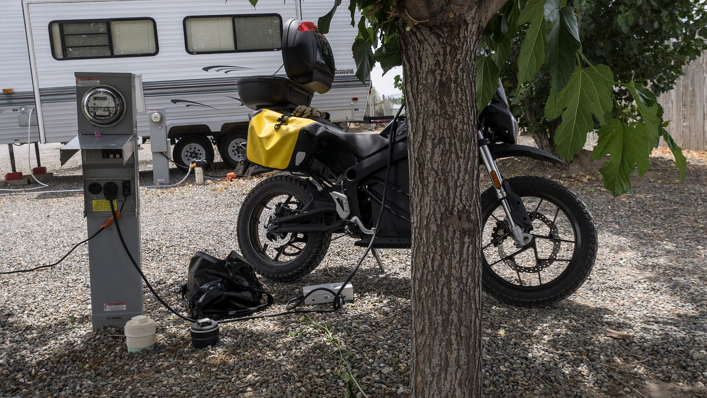 Photo of a Zero electric motorcycle plugged into an RV electrical panel and sitting in the shade of a tree. The bike rests upon a gravel parking area; there is a white travel trailer in the background. The topcase of the motorcycle is open and a thick black cord leads from the RV electrical panel to the motorcycle via an adapter box sitting upon the ground. The motorcycle is partially blocked by the trunk of a tree in the foreground. Some leaves from the tree are visible.