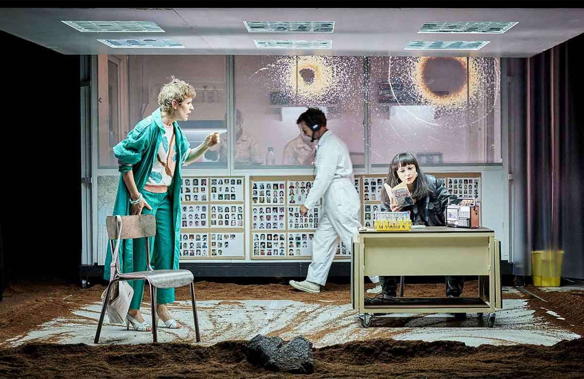 Scene from David Geselson's Neanderthal. A woman in a green rain coat talks to a woman in a leather jacket seated at a desk reading a book. There are photos of the faces of missing people pinned to the wall behind them. 