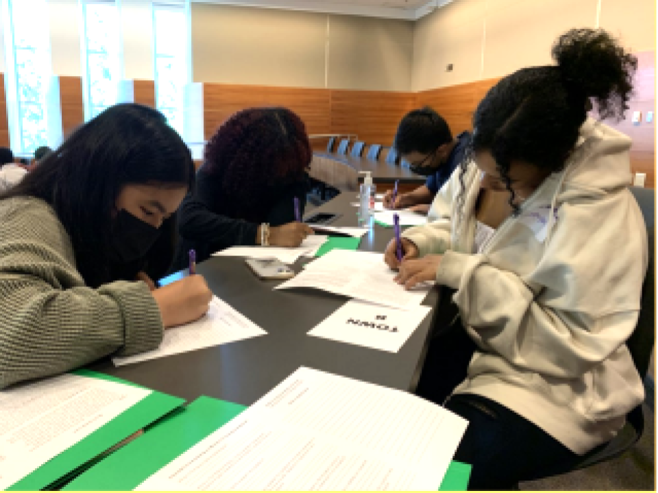Four high school aged youth of various racial backgrounds, masked, sitting at a long gray table in a building, two students on each side of the table. Students are gazing down at their work as they write with pens on white papers with green folders behind them.