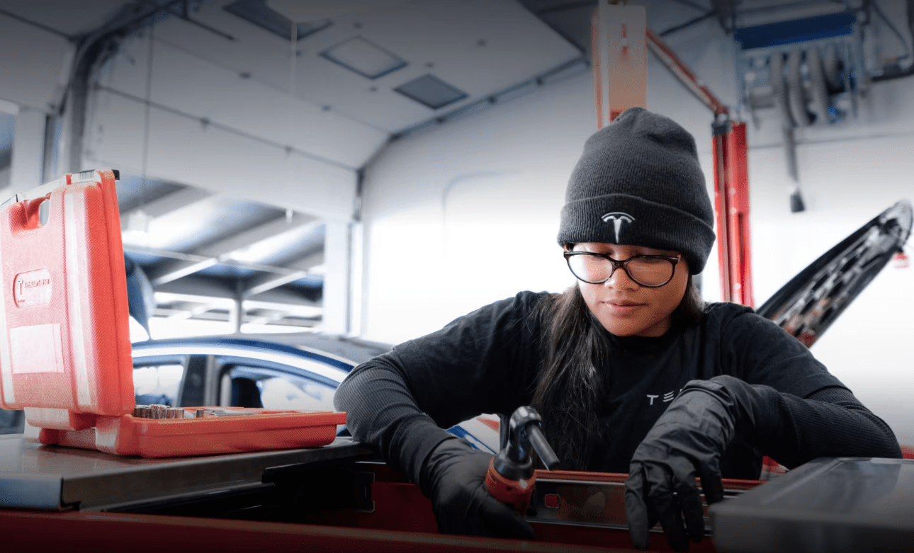 In a bustling garage setting, where green jobs are embraced, a person wearing a beanie and gloves skillfully uses a tool, with an electric car in the background and an open toolbox by their side.