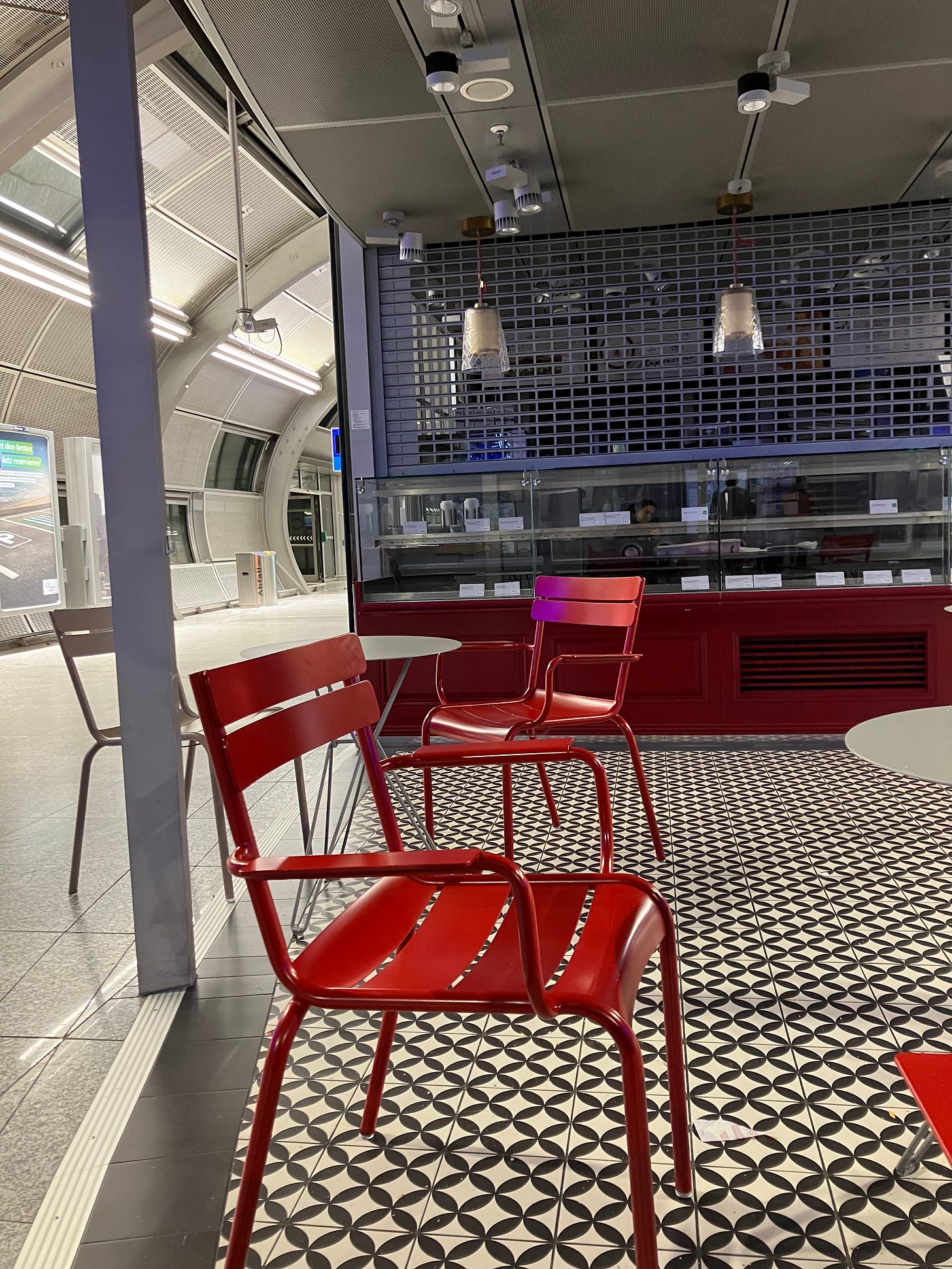 An airport corridor cafe with empty red chairs at empty round tables and the shutter down over the serving hatch.