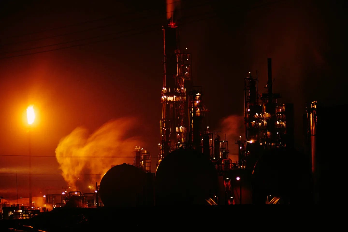 Smoke billows from an oil refinery in the Permian Basin at night, with a gas flare lighting up the scene.