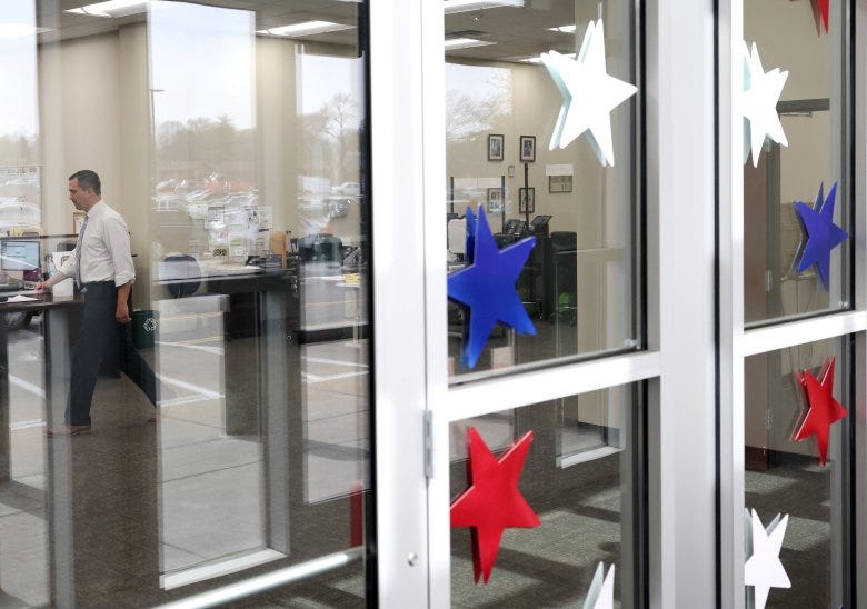 In the foreground are doors decorated with stars colored red, white and blue. The parking lot beyond reflects off the glass. Eric Fey is inside, walking past desks.