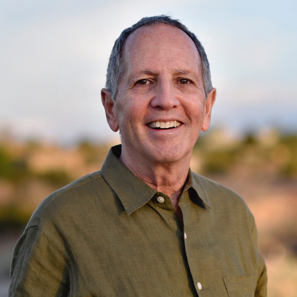 Alan Questel, A smiling man with short gray hair, radiating kindness, stands outdoors against a blurred natural background of greenery and a blue sky. He is wearing an olive green collared button-up shirt.