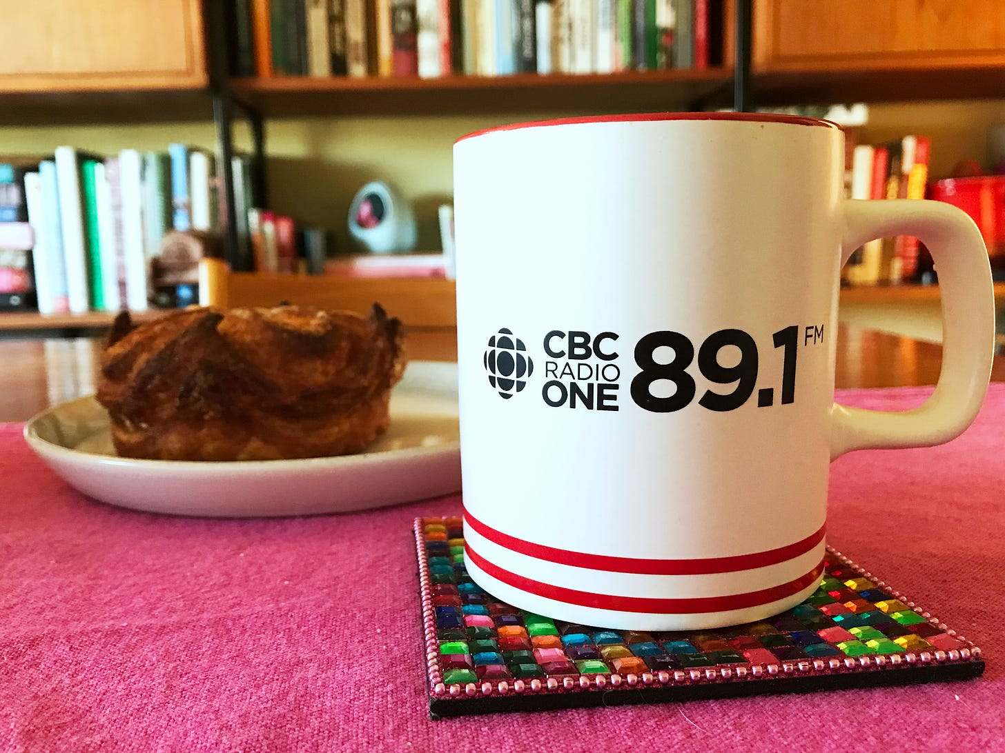 A photo of a CBC mug with the words "CBC Radio One 89.1 FM" on it. Behind it is a kouign-amman. They are sitting on a fuchsia table runner on my dining room table. Behind then are my bookcases, filled with cookbooks.