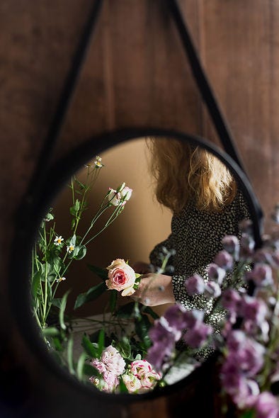 Picture of a round mirror reflecting partial image of a blonde haired women and flowers. 