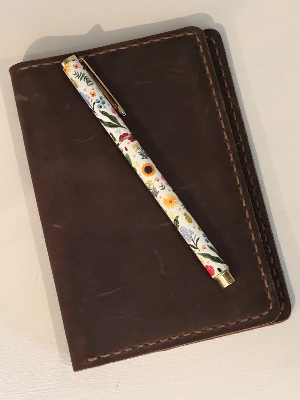 A little brown leather notebook cover with a small pen on top. The pen is white with illustrations of flowers 