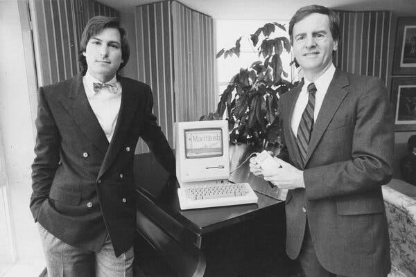 In a black-and-white photo, a man in a double breasted jacket and bow tie and a man in a gray suit and striped tie stand on either side of a computer that says “Macintosh” on its screen.