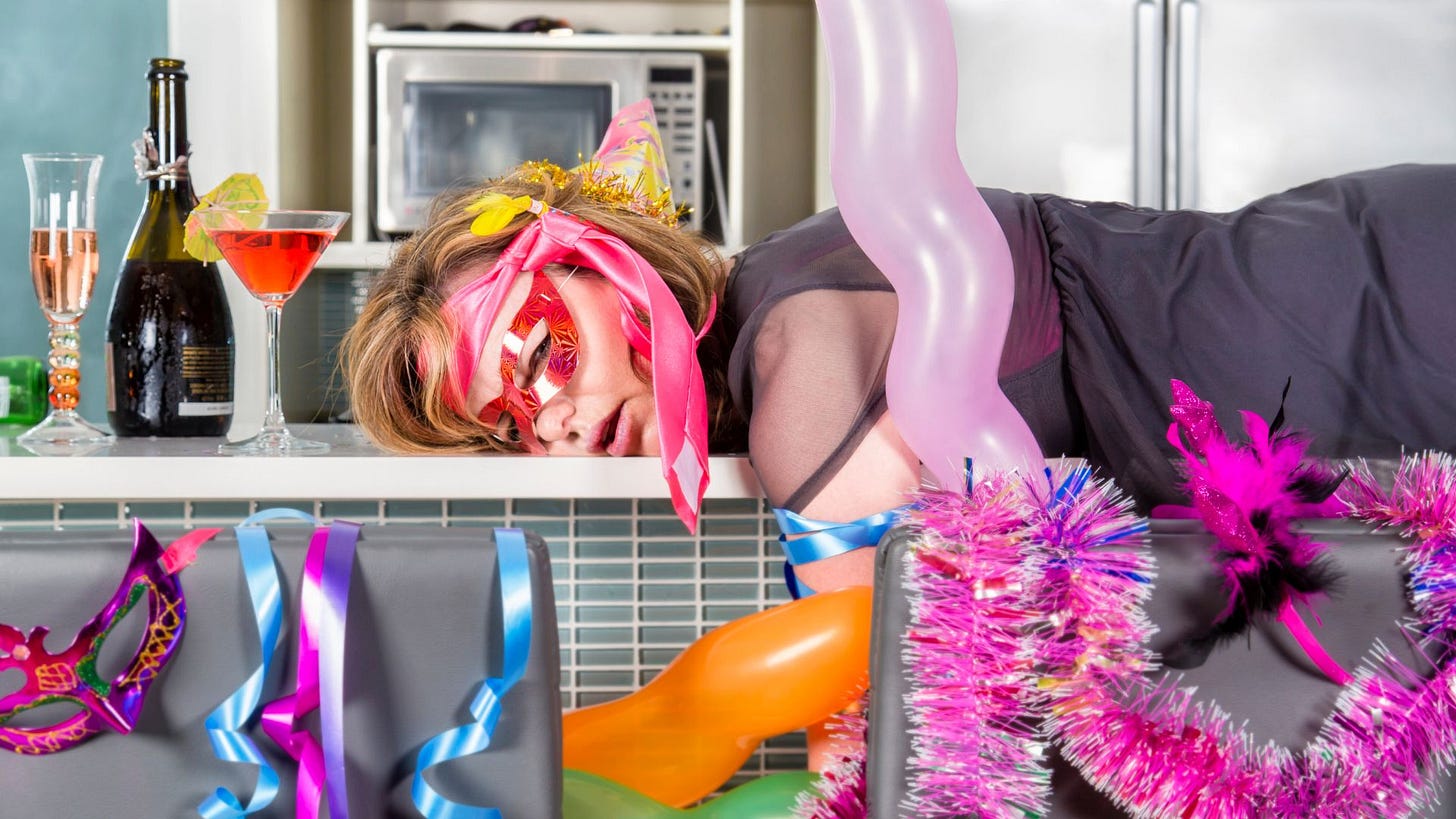 The photo of a young partied-out woman lying on the kitchen counter looking at the camera through an eye mask with bright balloons and ribbons hanging around, a champaign bottle and two stem glasses sitting at the woman’s head, and a white microwave in the background.